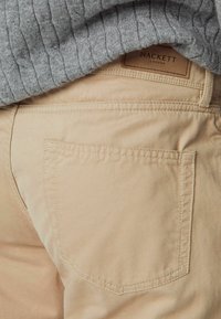 Beige bomuldsbukser med en baglomme, som viser en stikning. En grå ribbet sweater er delvist synlig over taljen.