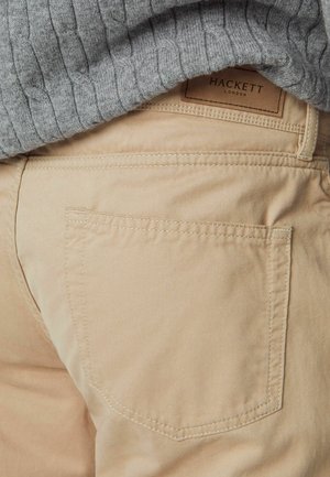 Beige katoenen broek met een achterzak, voorzien van een gestikte rand. Een grijze ribbeltrui is gedeeltelijk zichtbaar boven de tailleband.