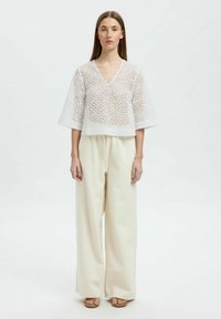 Hvid broderet top med brede ærmer, v-hals og åben arbejdsdesign. Parret med lyse beige vide bukser og flade sandaler.