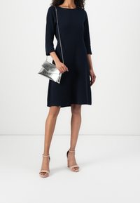 Tmavě modré šaty áčkového střihu s rukávy tři čtvrtě, doplněné o stříbrnou metalickou kabelku a tělové boty na podpatku s páskem kolem kotníku.