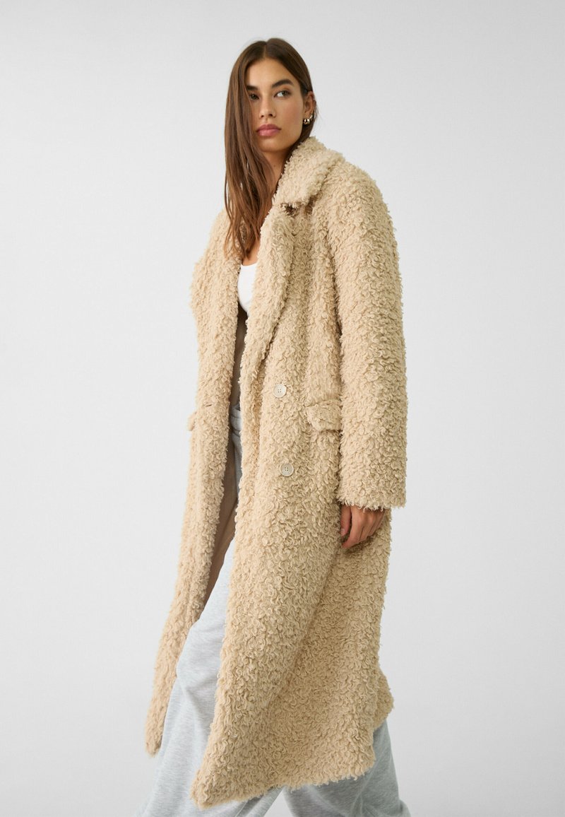 Kobieta w długim, beżowym, kręconym futrze teddy, nosząca białą bluzkę i szare spodnie, stojąca na tle jasnego, jednolitego tła.