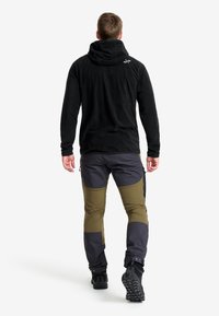 Schwarzer Fleece-Hoodie mit Kapuze, kombiniert mit grauen und olivgrünen Cargo-Hosen mit Reißverschlüssen und verstärkten Knien. Schwarze Wanderschuhe.