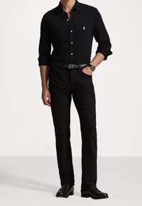 Zwarte lange mouwen blouse met knopen en kraag, met een wit logo op de borst. Gecombineerd met zwarte jeans en zwarte nette schoenen.