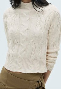 Suéter de punto cableado en color crema, con un cuello alto de tipo barco, dobladillo acanalado y un patrón texturizado que presenta trenzas y giros elevados en la tela.