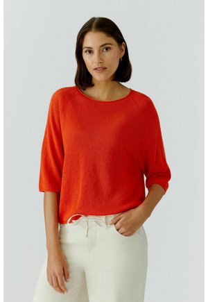 Frau mit schulterlangen dunklen Haaren, die ein leuchtend orangefarbenes Strickoberteil und cremeweiße Hosen trägt, steht mit einer Hand in der Tasche.