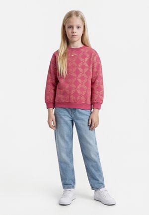 Jeune fille aux longs cheveux blonds, portant un sweat-shirt rose à motifs, un jean bleu clair et des baskets blanches, debout sur un fond blanc.
