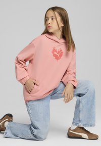 Sudadera rosa con un logo de corazón en forma de llama, con mangas largas y un bolsillo canguro, combinada con jeans de color azul claro y zapatillas marrones.