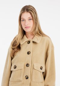 Beige fleecejakke med krave, frontlukning med knapper og to brystlommer med klap. Blød tekstur med en afslappet pasform.