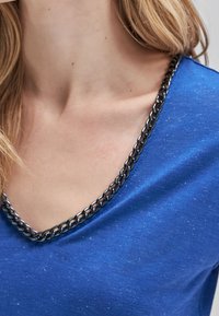 Niebieska bluzka w serek z metalicznym akcentem wzdłuż dekoltu, wykonana z miękkiego materiału z delikatnym połyskiem.