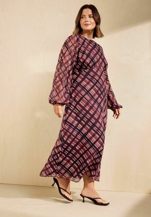 Woman wearing a long, purple and black patterned dress with sheer balloon sleeves and black open-toe heeled sandals, standing against a neutral wall.