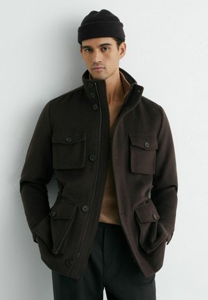 Mees, kes kannab tumepruuni nööpide ja tõmblukuga jope nelja taskuga, musta mütsi ja musti pükse, seisab valge seina ääres.