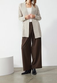 Krémový pletený svetr s kabelovým designem, otevřený vpředu, s žebrovanými manžetami, kombinovaný s hnědými kalhotami širokého střihu s vysokým pasem a černými botami na vysokém podpatku.