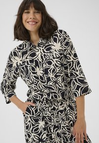 Black and cream patterned shirt featuring a floral design, short sleeves, a button-up front, and two chest pockets. Textured fabric.
