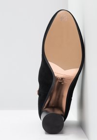 Chaussure à talon en daim noir montrée par la semelle, dotée d’un talon rond et d’une semelle lisse marron clair avec les détails de la taille et du logo.
