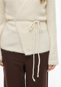 Krēmkrāsas džemperis ar teksturētu adījuma materiālu. Tam ir saite jostas vietā un garas piedurknes ar atvērtu V veida kakla izgriezumu.