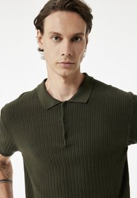 Mörkgrön ribbad polo-tröja med klassisk krage, korta ärmar och tre knappar framtill. Slät textur med en åtsittande design.