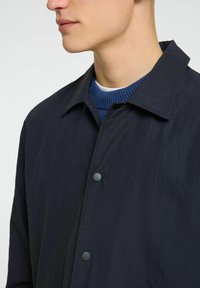 Giacca navy con finitura opaca e colletto a punte, dotata di bottoni a pressione neri e sovrapposta a un maglione blu a coste sottostante.