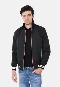 Giubbotto bomber nero con colletto alto, dettagli bianchi e rossi, tasche con zip e polsini a costine, indossato sopra una maglietta nera e jeans blu.