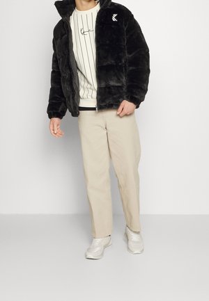 Personne portant une veste en fausse fourrure noire, un pull rayé blanc, un pantalon beige et des baskets blanches, debout devant un fond uni.