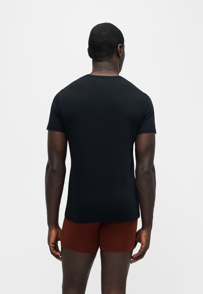 Homme debout, de dos, portant un t-shirt noir près du corps et un short rouge foncé sur un fond clair uni.
