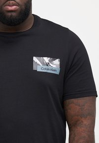 Melnā kokvilnas T-kreklā ir neliels grafiskais kabatas izdrukas motīvs ar vienkrāsainu druku un zilu aksentu, kurā ir uzraksts "Calvin Klein."