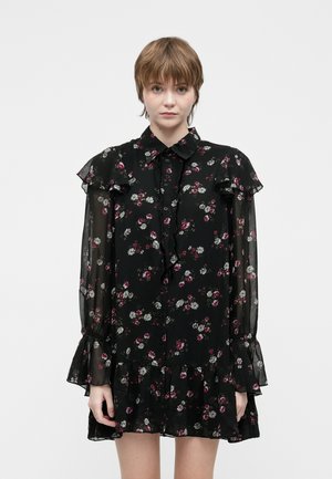 Jonge vrouw met kort haar, gekleed in een zwart bloemetjes doorzichtige lange mouwen mini-jurkje met ruffles bij de schouders, manchet en zoom.