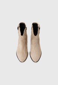 Botine din suede bej, cu vârf ascuțit, guler cu margini ondulate și închidere cu fermoar pe lateral. Textură netedă și design minimalist.