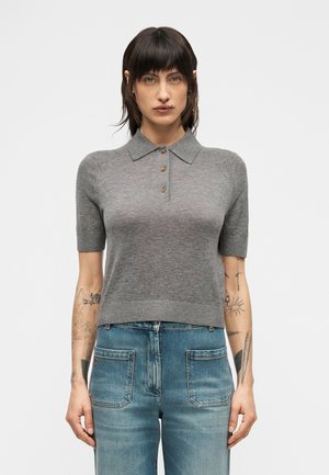 Femme aux cheveux courts et foncés, vêtue d'un polo gris à manches courtes et d'un jean bleu taille haute, debout devant un fond blanc uni.