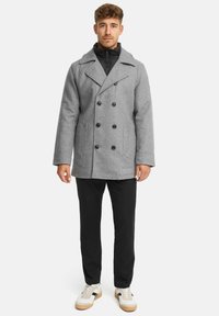 Grå uld peacoat med dobbeltradede knapper og klippet krave, lagd over en sort zip-up jakke. Sorte bukser og hvide sneakers fuldender outfittet.