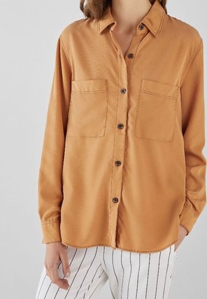 Woman wearing a long-sleeve tan button-up shirt with two chest pockets, paired with white pants featuring black vertical stripes.