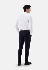 Weißes Langarmhemd mit einem geraden Kragen, kombiniert mit navyblauen Hosen. Der Hintergrund ist schlicht weiß und hebt die Passform und das Design des Outfits hervor.