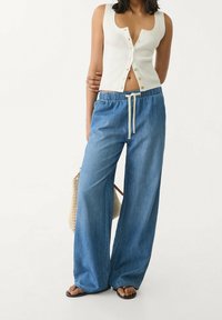 Kvinna i ärmlös vit knäppad crop top, lösa blå denimbyxor med dragsko, svarta sandaler och håller en vävd väska.