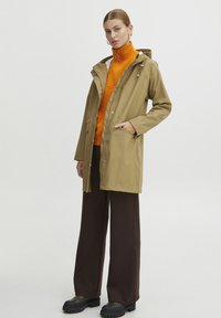 Vattentålig beige regnrock med huva, sidofickor och tryckknappar över en orange tröja och bruna vida byxor. Kraftiga svarta stövlar.