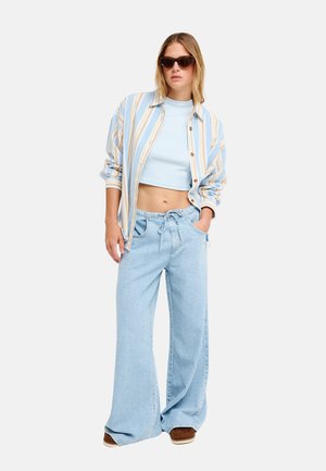 Femme portant de grandes lunettes de soleil, une chemise rayée ouverte, un crop top bleu clair, un jean à jambes larges et des chaussures marron sur un fond uni.
