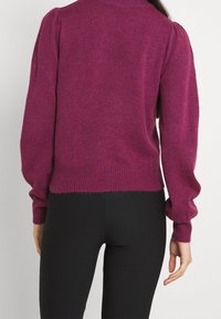 Magenta dziergany sweter z wysokim kołnierzem, bufkami na rękawach i ściągaczem na dole. Tekstura jest miękka i lekko puszysta. Noszony z czarnymi spodniami.