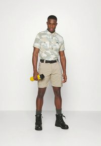 Kortermet lys grå kamuflasjeskjorte, beige shorts, svart belte, svarte kampstøvler, holder en gul vannflaske.