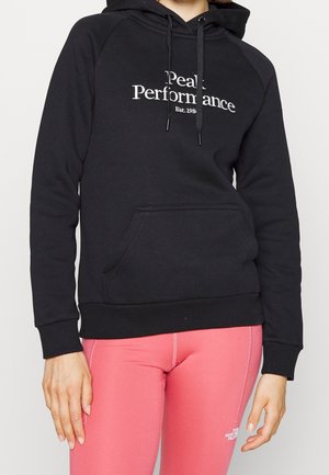Osoba v černé mikině s kapucí s nápisem "Peak Performance Est. 1986" a růžovými legínami s logem The North Face na stehně.