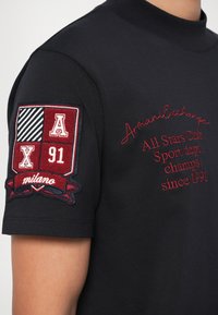 Maglietta nera con scollo tondo, dotata di un logo ricamato rosso e una toppa con motivi geometrici in bordeaux, bianco e blu sulla manica.