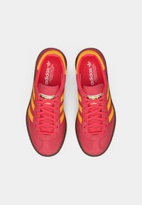 Κόκκινα αθλητικά παπούτσια Adidas με κίτρινες ρίγες και κορδόνια, φωτογραφημένα από ψηλά σε λευκό απλό φόντο.