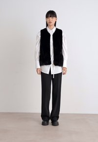 Schwarze Kunstpelz-Weste über einem weißen Hemd mit Knöpfen, kombiniert mit lockeren schwarzen Hosen und schwarzen Schuhen, vor einem schlichten weißen Hintergrund.