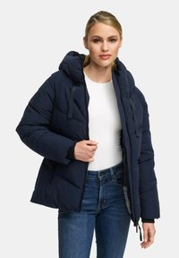 Vrouw draagt een marineblauwe gewatteerde jas met capuchon, een wit shirt en blauwe jeans, terwijl ze de rits van de jas vasthoudt.