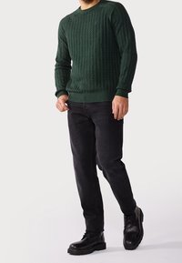 Maglione verde lavorato a maglia con motivo a trama, abbinato a jeans neri e stivali neri con lacci, evidenziato da un design aderente e polsini a coste.