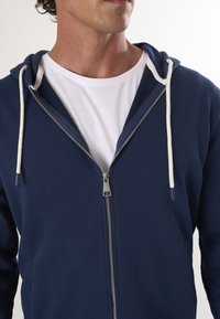 Sudadera con cremallera de color azul marino y cordones blancos. Presenta una cremallera plateada, bolsillos tipo canguro y un tejido suave y texturizado. Camiseta blanca debajo.