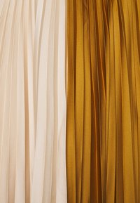 Pionowe panele z plisowanej tkaniny ułożone obok siebie, jeden w jasnym beżu, drugi w musztardowej żółci, z miękkimi, równymi fałdami.