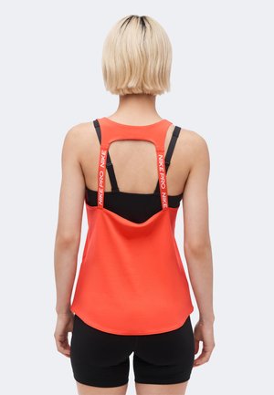 Žena s krátkymi blond vlasmi nosí červené tielko Nike Pro cez čiernu športovú podprsenku a čierne šortky, zobrazená zo zadnej strany na bielom pozadí.