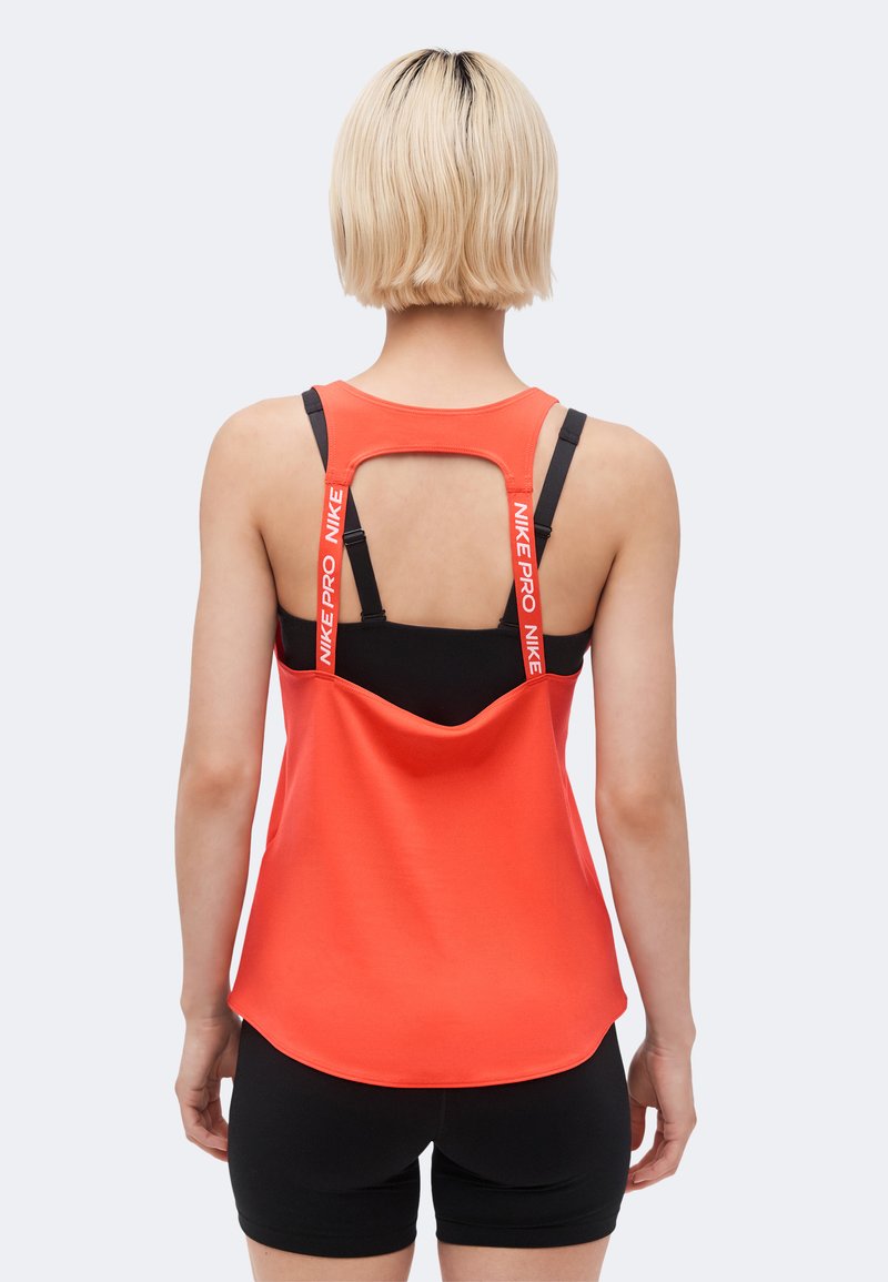 Γυναίκα με κοντά ξανθά μαλλιά φοράει κόκκινο αμάνικο Nike Pro πάνω από μαύρο αθλητικό σουτιέν και μαύρα σορτς, εμφανίζεται από πίσω σε λευκό φόντο.