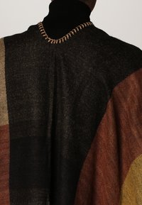 Person wearing a multi-colored knit poncho with black, brown, tan, and mustard yellow sections over a black turtleneck.