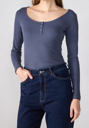 Vrouw draagt een aansluitend geribbeld grijs langarmtopje met een ronde halslijn en drie knopen, gecombineerd met een hooggetailleerde donkerblauwe spijkerbroek.