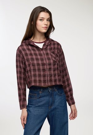 Camicia a quadri tagliata in un bordeaux scuro e rosa, con cappuccio, bottoni frontali e maniche lunghe. Abbinata a jeans blu a vita alta.