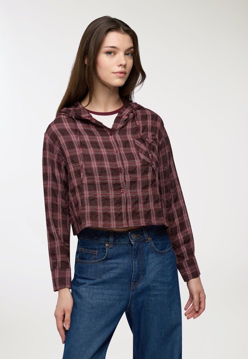 Camicia a quadri tagliata in un bordeaux scuro e rosa, con cappuccio, bottoni frontali e maniche lunghe. Abbinata a jeans blu a vita alta.